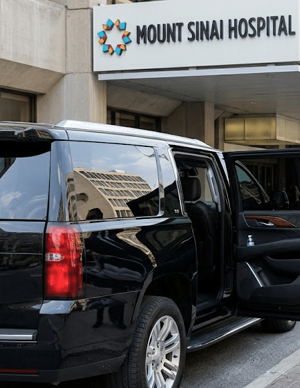 A professional chauffeur in a suit standing by an open black SUV door at the Mount Sinai Hospital entrance in Toronto, holding a sanitized infant car seat.