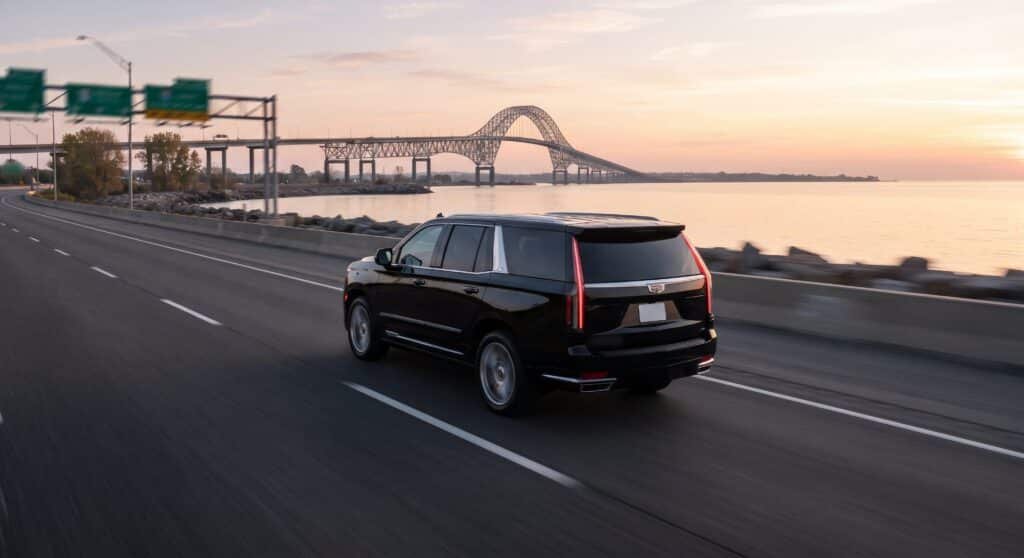 Black Cadillac Escalade on a lakeside highway at sunrise with a steel arch bridge behind, flat-rate car service from Hamilton to Toronto